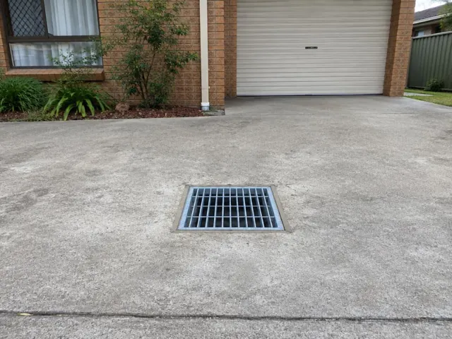 Concrete driveway with a square metal drain grate in front of a garage in Metford
