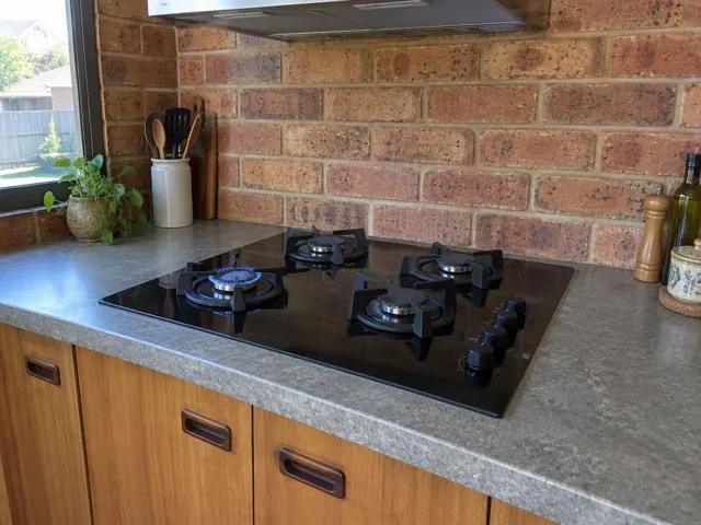 Black gas cooktop set into a grey benchtop with brick backsplash and timber cabinets in Thornton