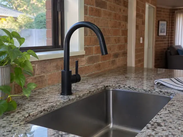 Black kitchen mixer tap over stainless sink with granite benchtop and brick wall in Metford